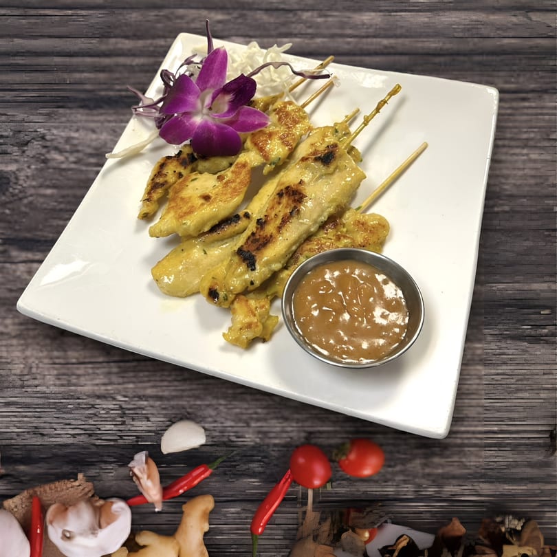 Grilled chicken satay skewers with peanut sauce and an orchid garnish on a white plate.