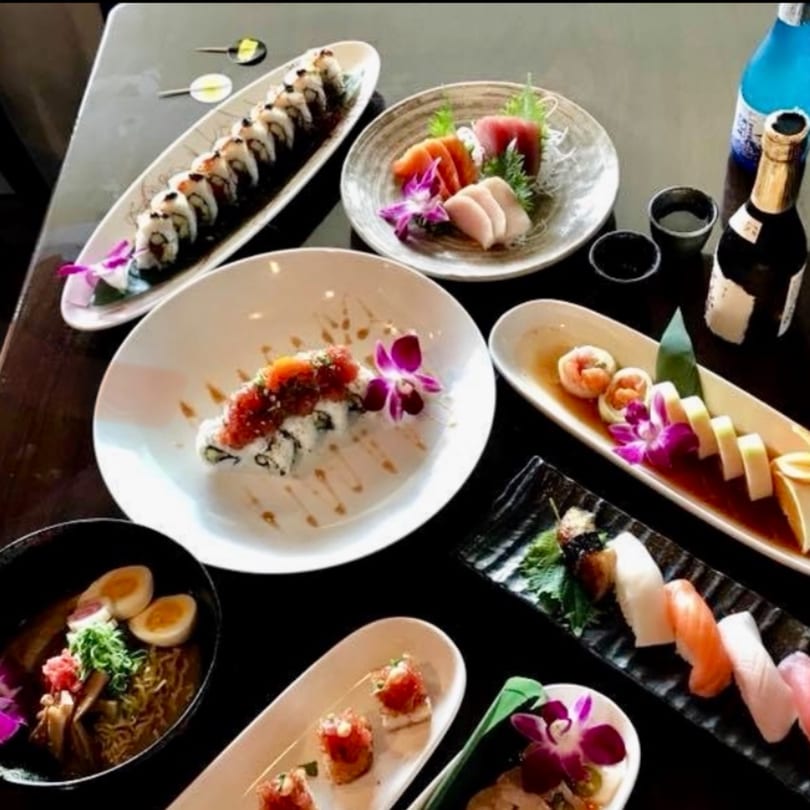 A table filled with various sushi rolls, sashimi, ramen, and sake bottles at Kenzo Thai & Sushi.