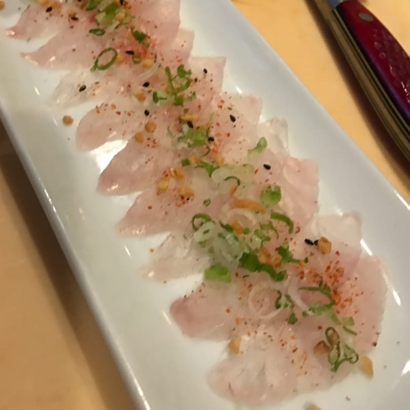 Thinly sliced white fish sashimi, garnished with green onions, spices, and sesame seeds on a white plate.