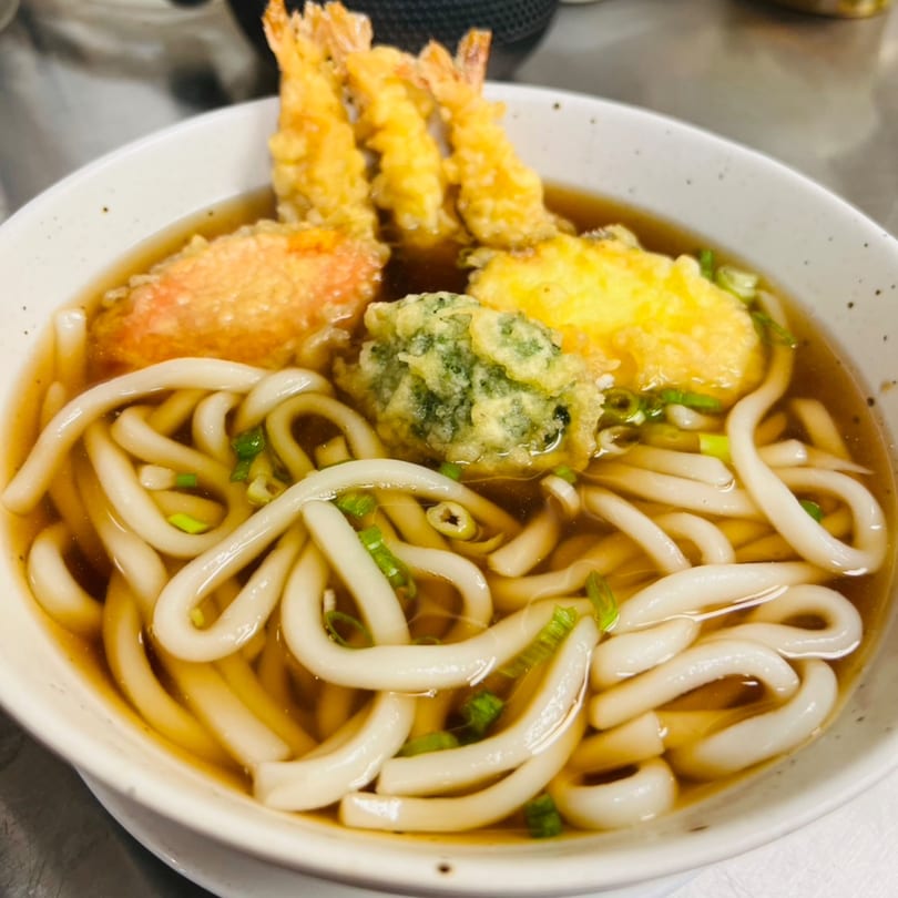A bowl of udon noodles with broth, topped with tempura shrimp and vegetables.
