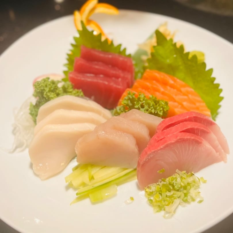 A white plate with various sashimi slices, including tuna, salmon, and white fish, garnished with greens.