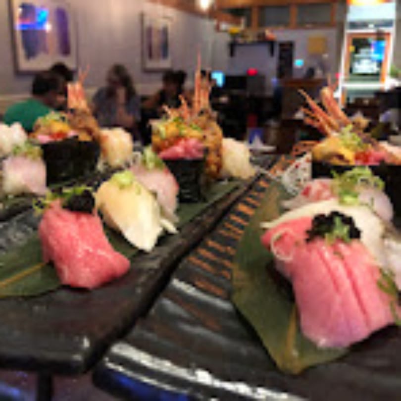 A close-up of various sushi and sashimi dishes on dark plates, with blurred diners in the background.