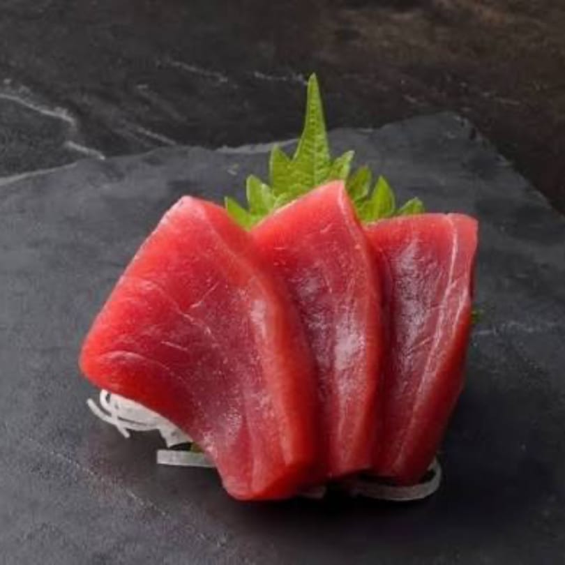 Three slices of vibrant red tuna sashimi, garnished with green leaves, on a dark slate plate.