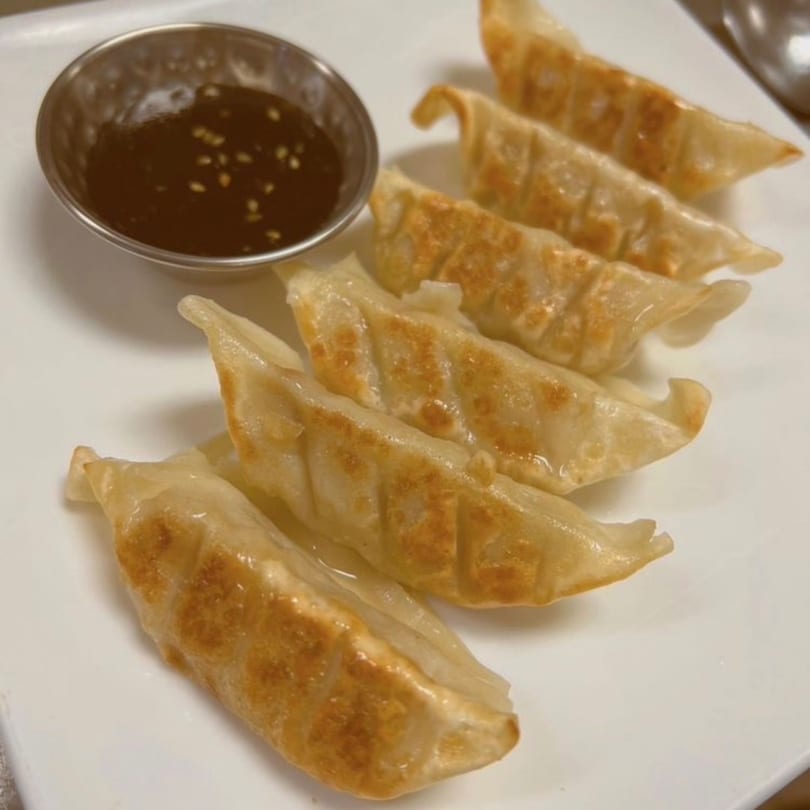Six golden-brown gyoza dumplings with a side of dipping sauce on a white plate.